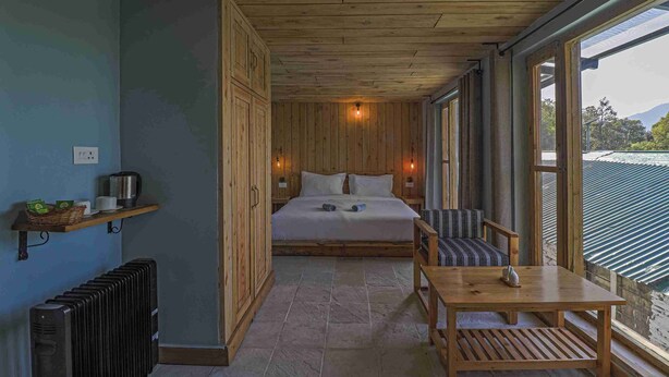 Una habitación con paneles de madera, que incluye una cama, una mesa, sillas, un radiador y una estantería con objetos.