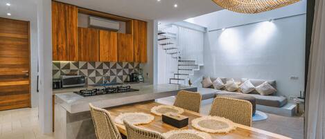 A modern kitchen with wooden cabinets, a central island with chairs, and a staircase leading to a living area with a sofa and cushions.