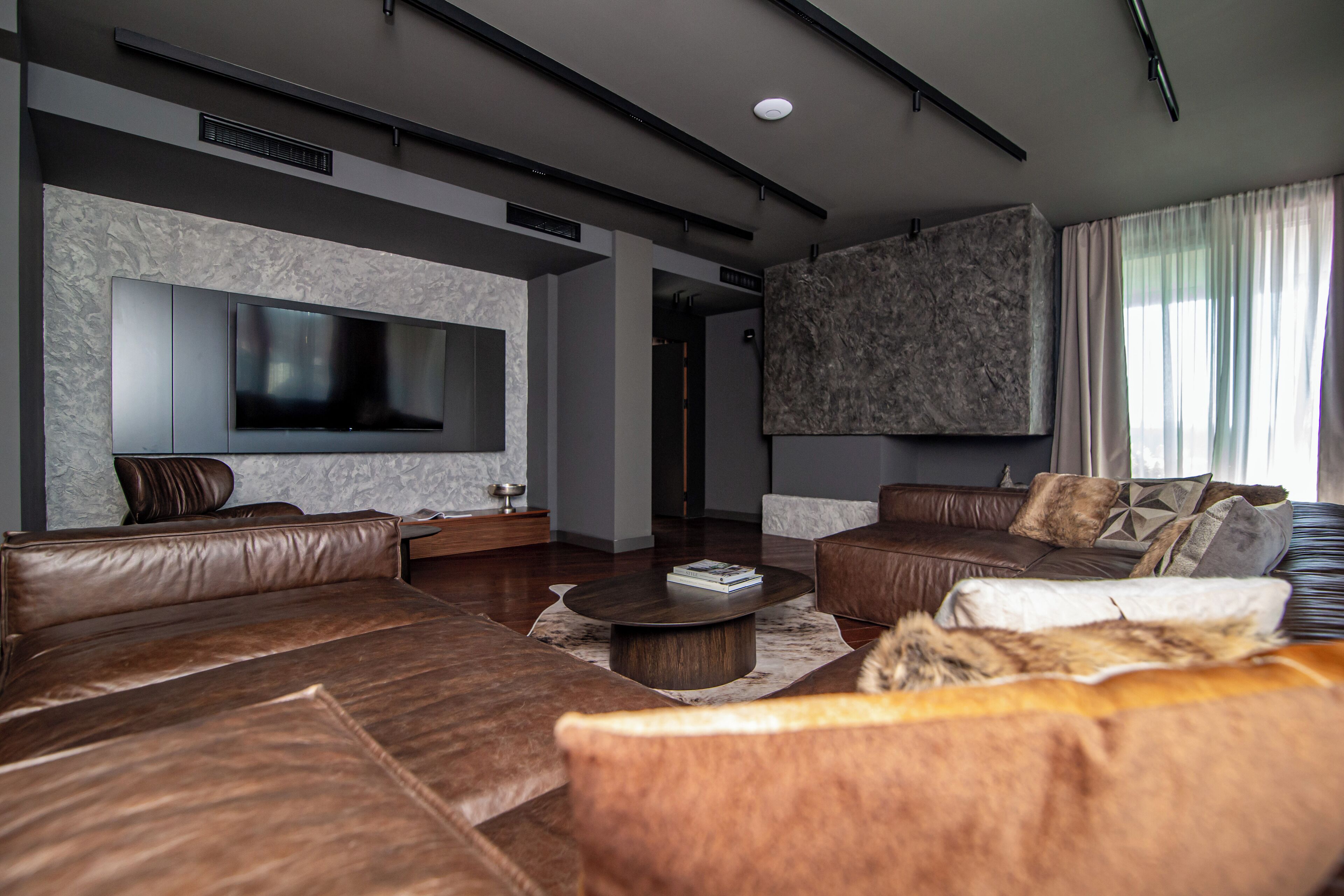 A modern living room with leather sofas, a flat-screen TV mounted on the wall, and a large window with curtains.