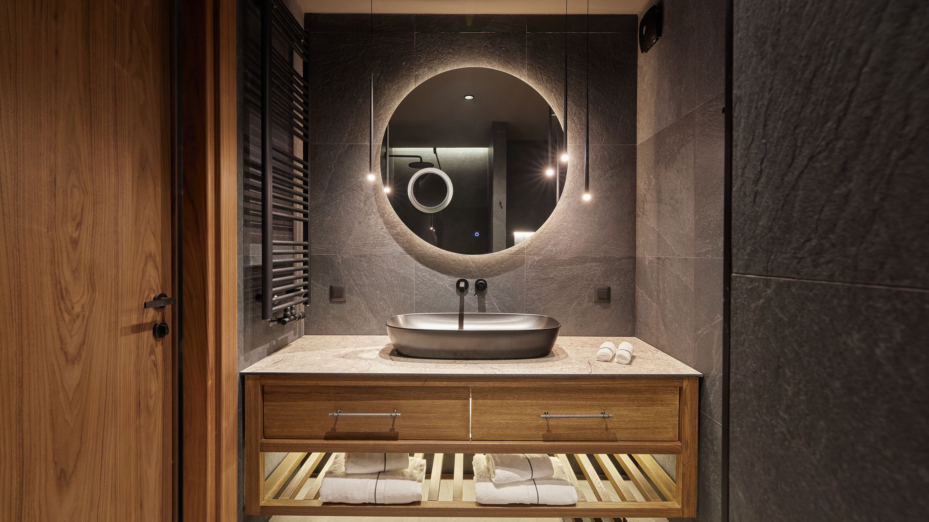 A modern bathroom with a round mirror, a sink, and a wooden vanity with a towel rack.