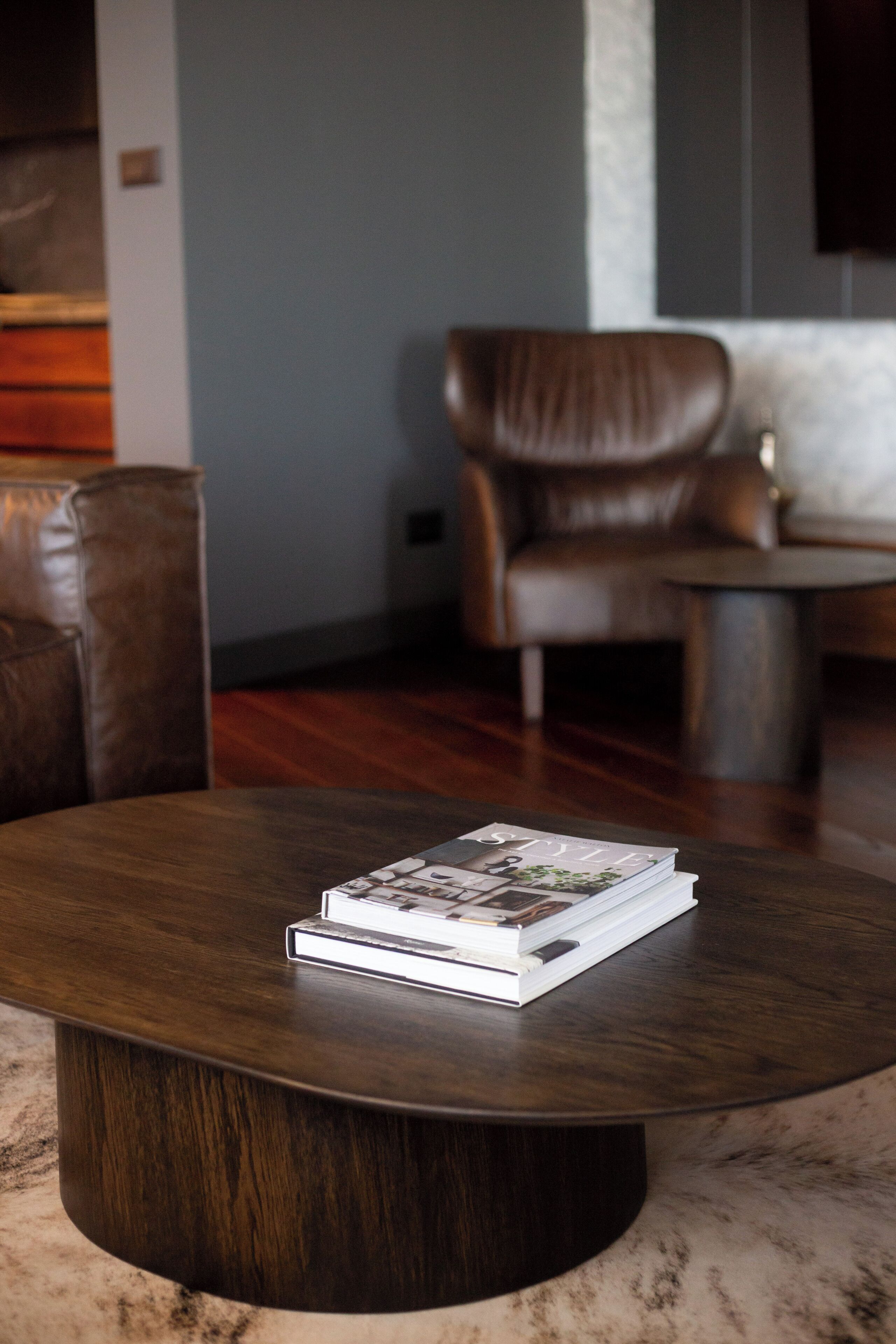 Ein runder Holztisch mit einer Zeitschrift in der Mitte, ein brauner Ledersessel und ein weißes Sofa im Hintergrund.