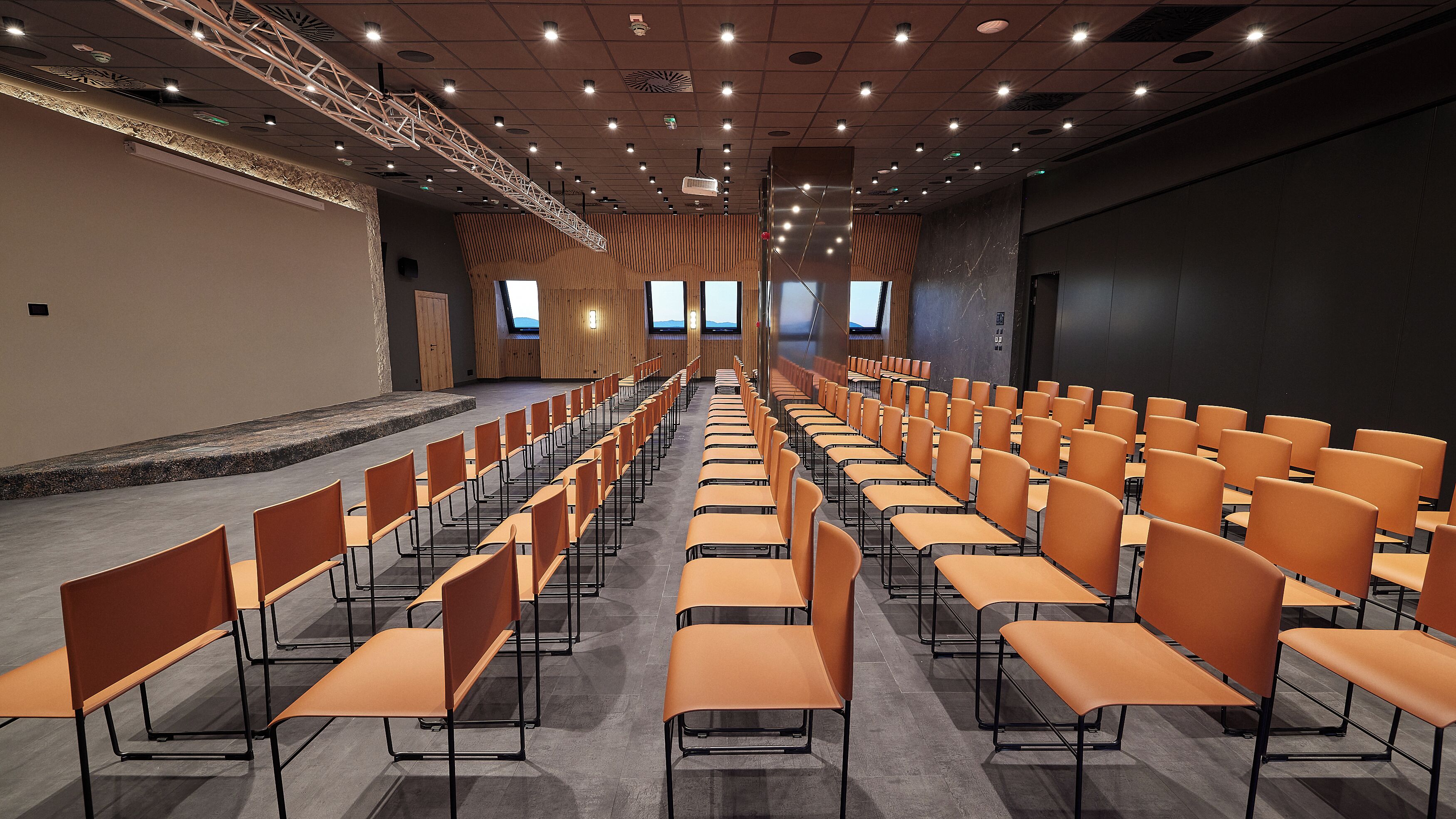 A conference room with rows of orange chairs, a stage, and a high ceiling with recessed lighting.