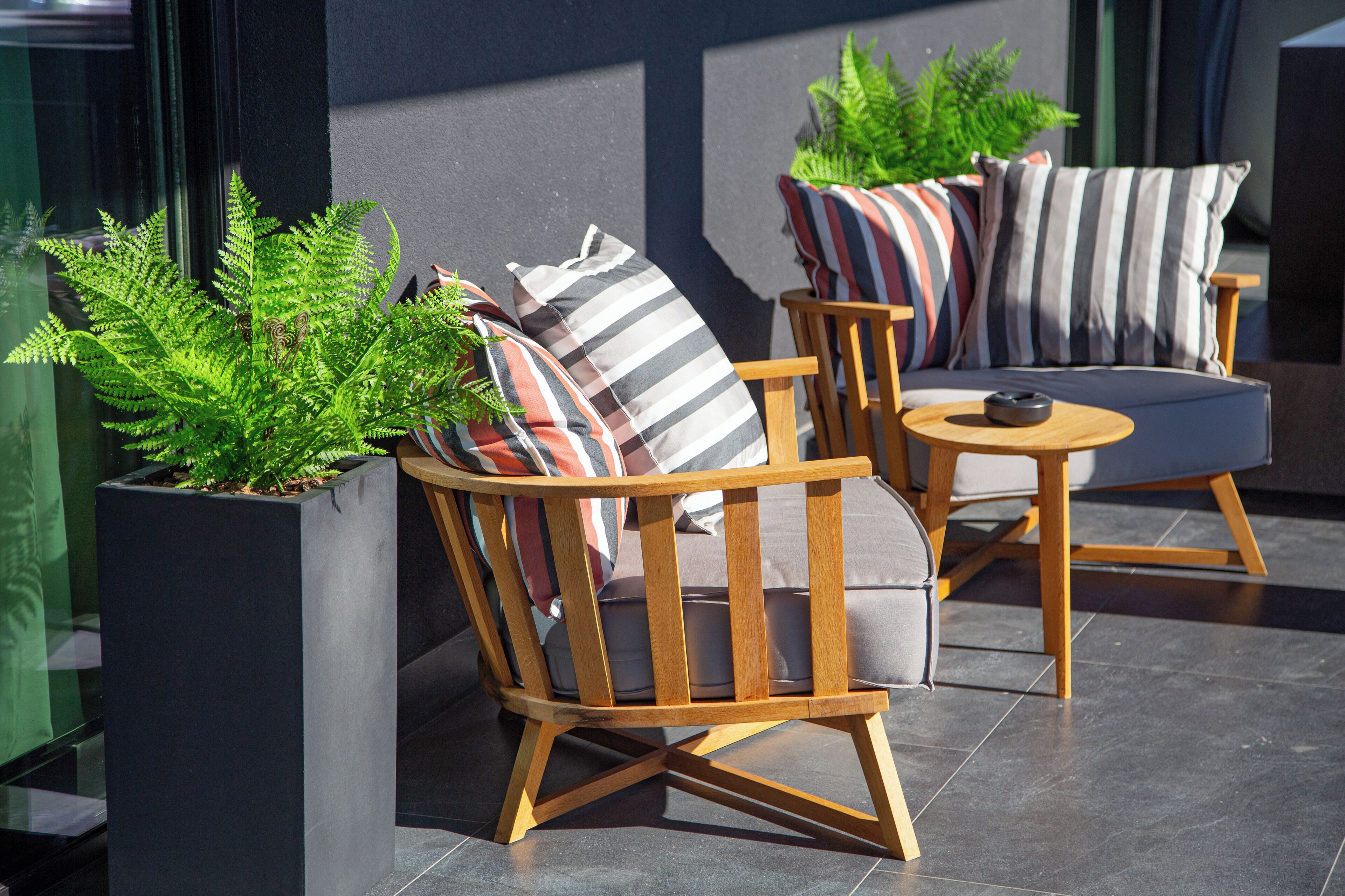 Un espacio al aire libre moderno con sillas de madera y una mesa redonda, rodeado de plantas en macetas.