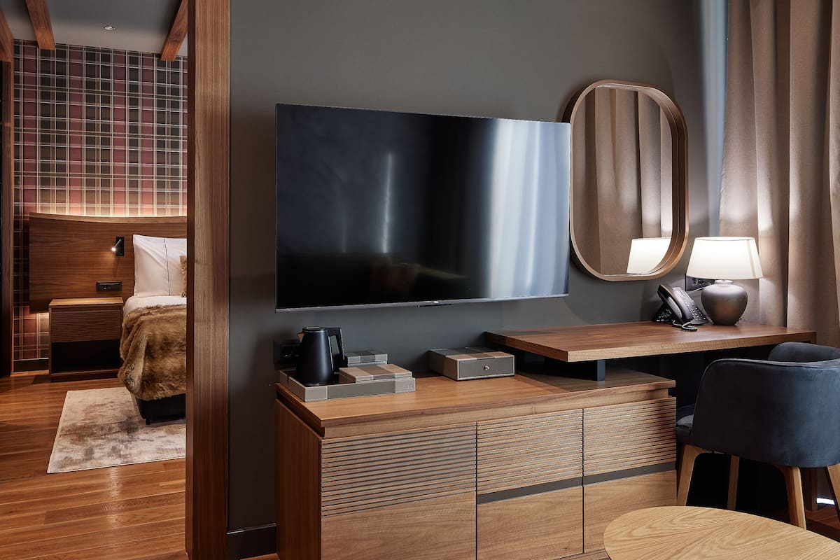 A hotel room with a large flat-screen TV, a bedside table with a lamp, a mirror, and a wooden cabinet.