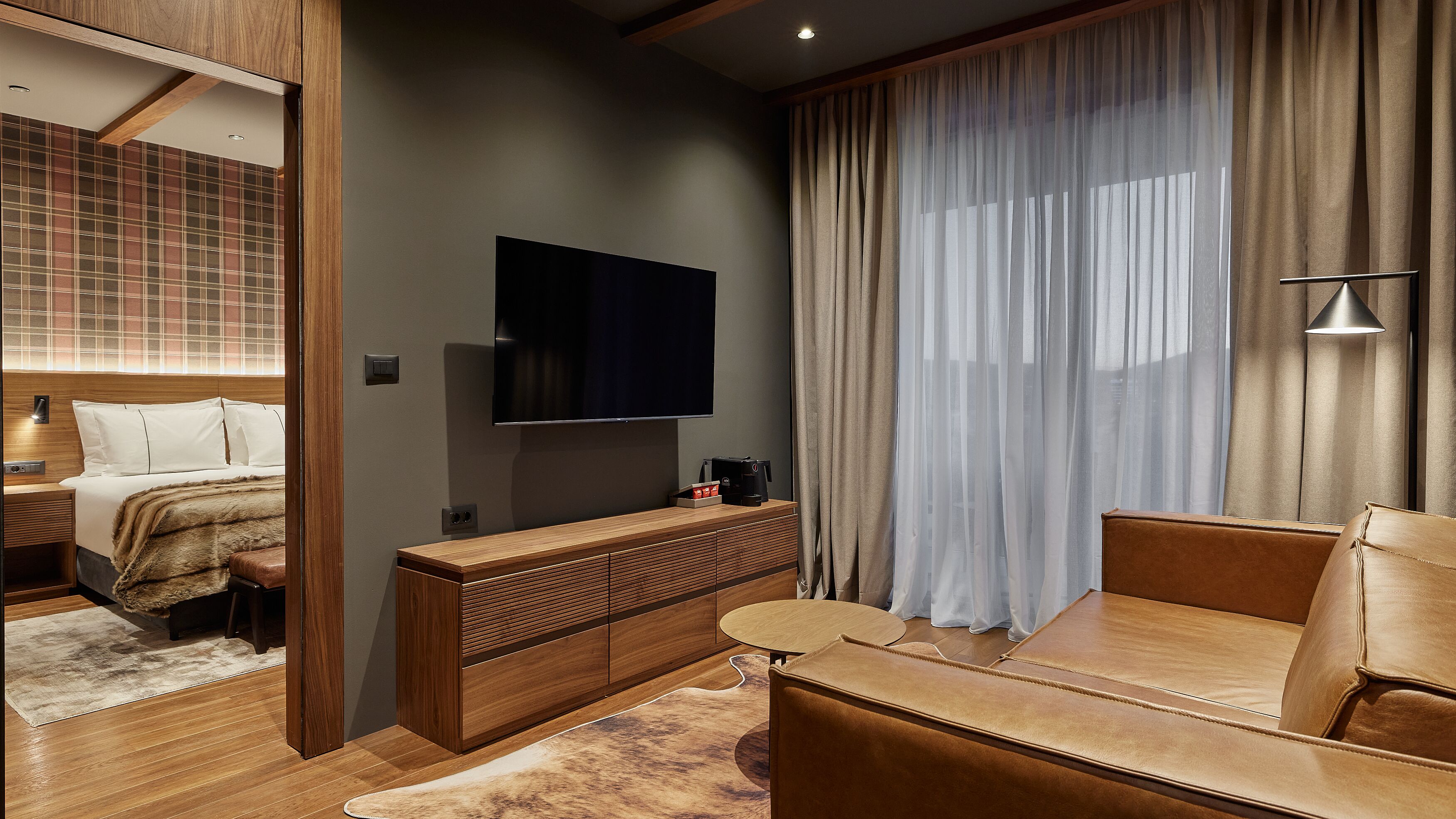 A modern hotel room with a large flat-screen TV, a wooden console table, a leather sofa, and a large window with sheer curtains.