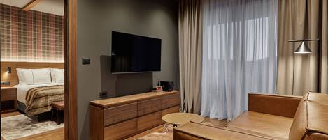 A modern hotel room with a large flat-screen TV, a wooden console table, a leather sofa, and a large window with sheer curtains.