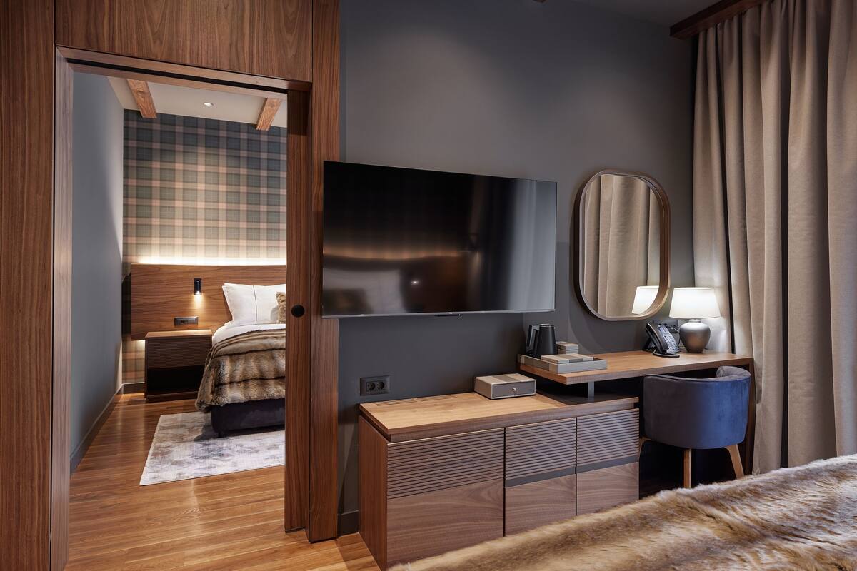A modern hotel room with a wooden wardrobe, a flat-screen TV, a bedside table with a lamp, a mirror, and a fur throw on the bed.