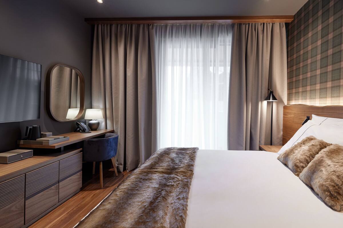 A hotel room with a large bed, a wooden desk, a mirror, a lamp, and a view of the outside through sheer curtains.