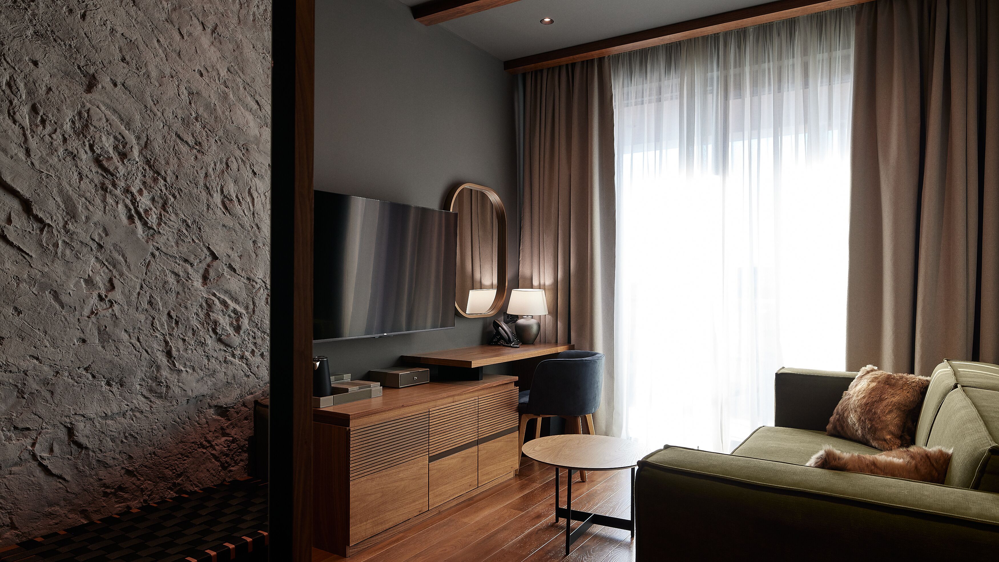 Une chambre d’hôtel moderne avec un mur à la texture de pierre, une console en bois, un téléviseur à écran plat, une table ronde et un canapé vert agrémenté d’un plaid en fausse fourrure.