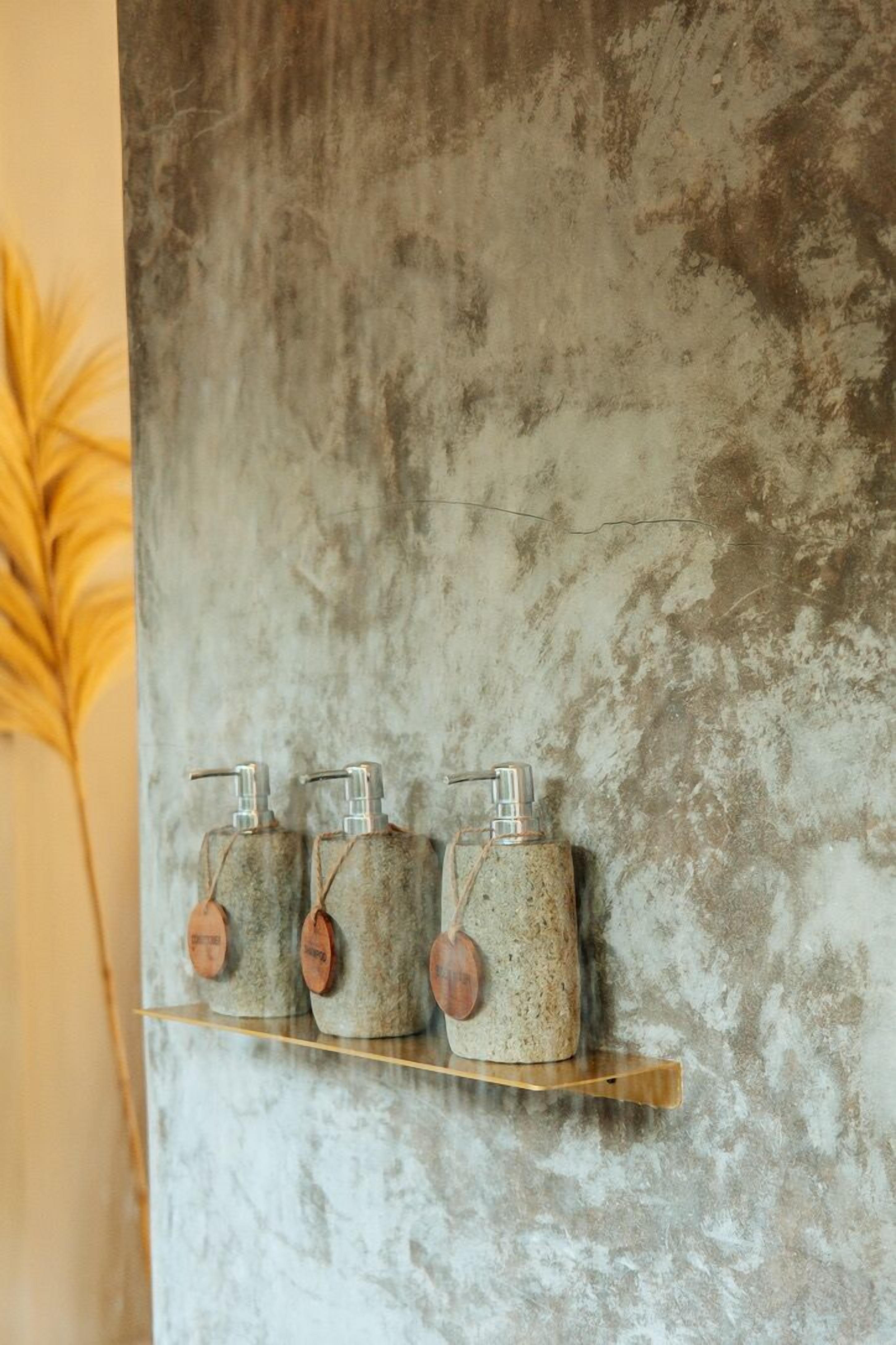 A wall-mounted shelf with three rustic, textured containers, each with a metal pump dispenser and a wooden tag.