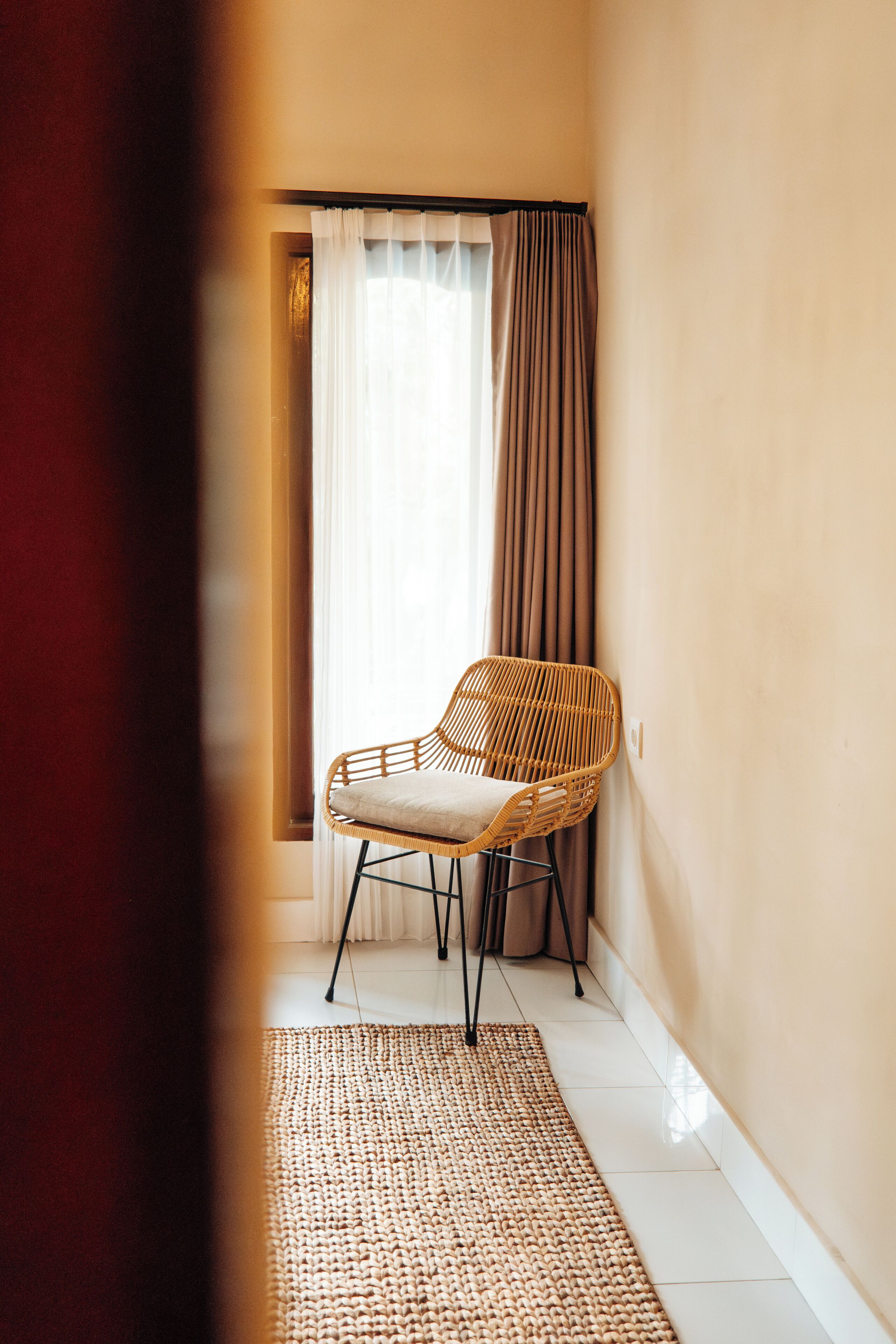 A narrow hallway with a wicker chair, a white rug, and a window with sheer curtains.