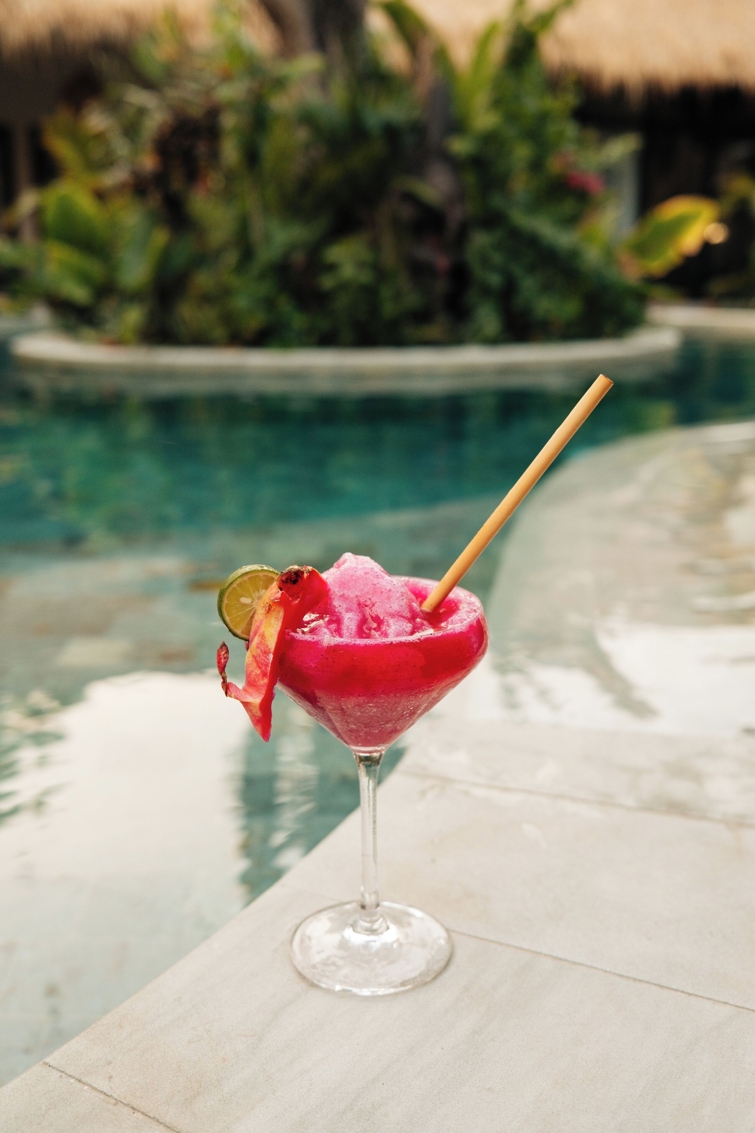 A pink cocktail with a lime garnish and a straw, placed on a table by a pool with a thatched roof in the background.
