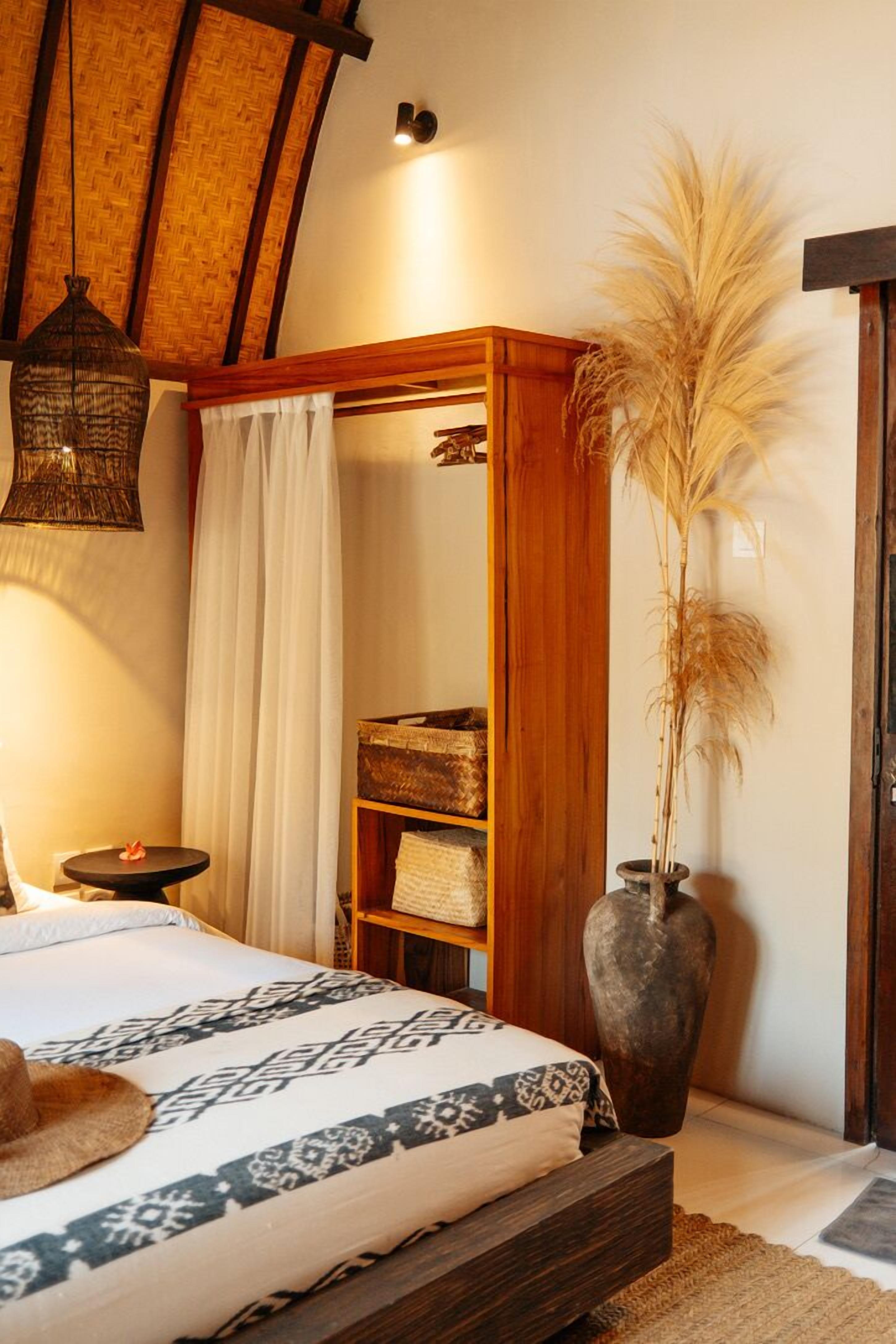 A bedroom with a large bed, a wooden wardrobe, a small table with a candle, and a tall vase with dried plants.