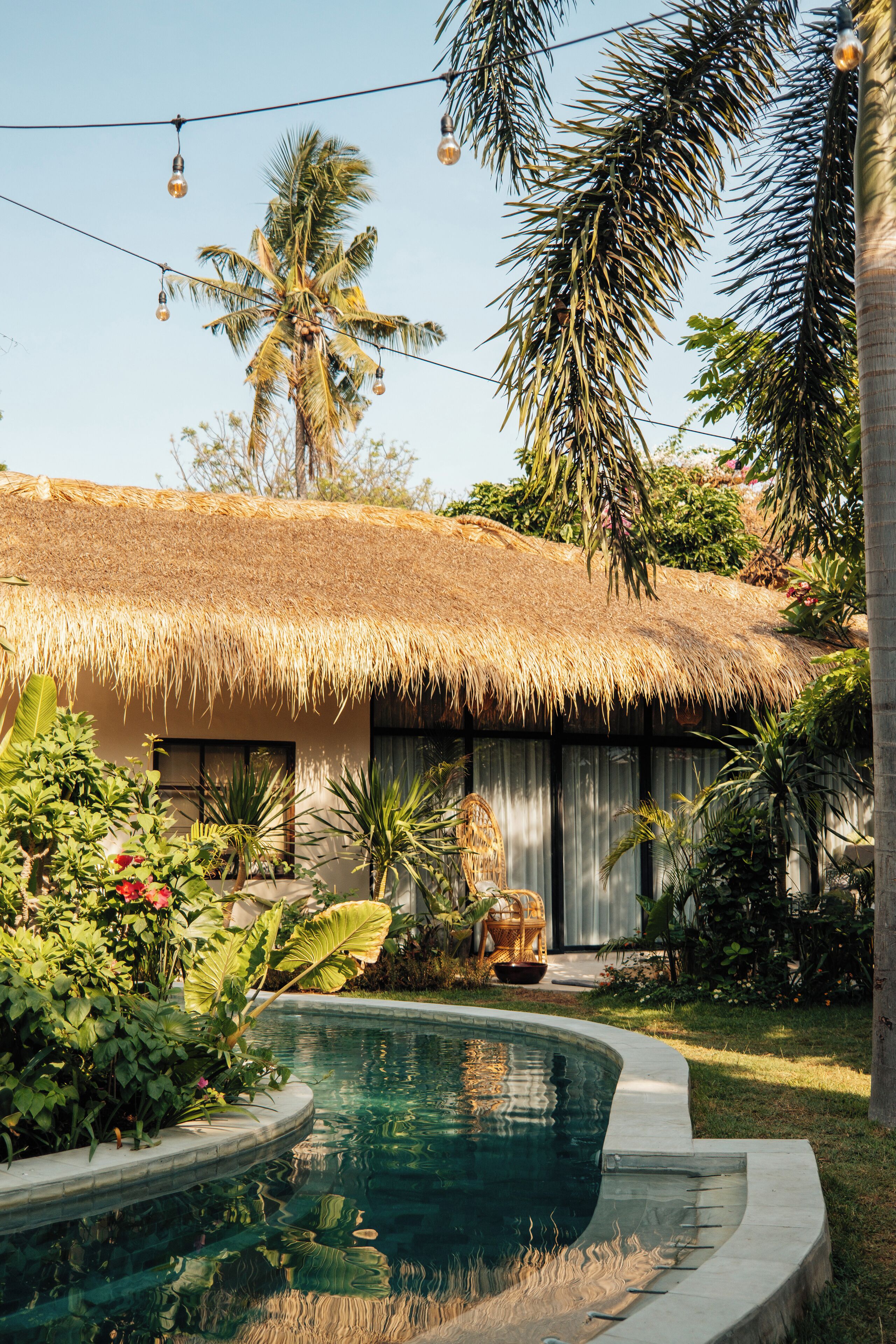 A poolside area with a thatched-roof structure, surrounded by lush greenery and a clear pool.