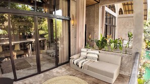 A modern interior with a glass door leading to an outdoor seating area, a sofa with patterned cushions, and a variety of potted plants.