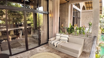 A modern interior with a glass door leading to an outdoor seating area, a sofa with patterned cushions, and a variety of potted plants.