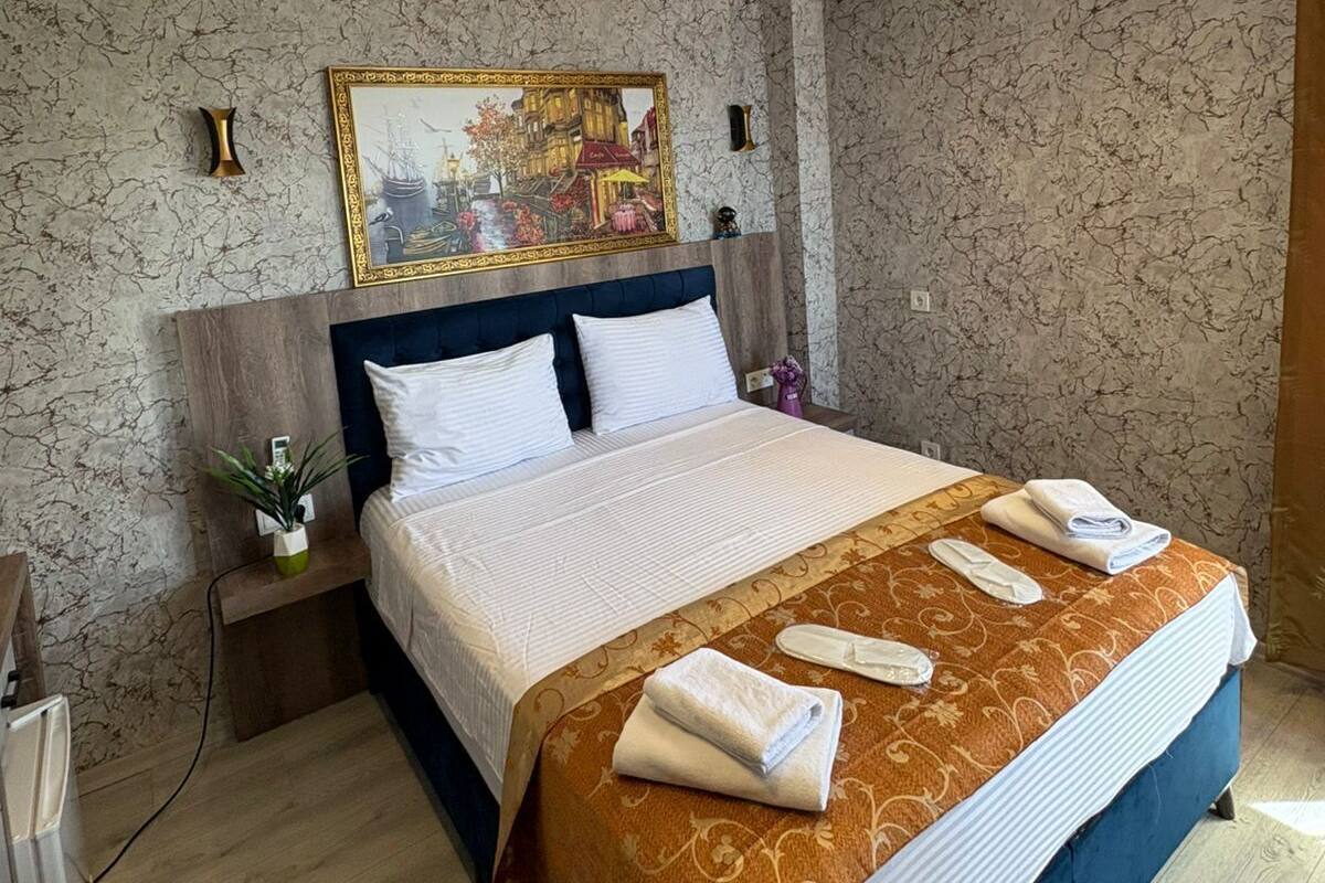 Una habitación de hotel con una cama grande, cabecera, un cuadro en la pared y una mesita de noche con una lámpara.
