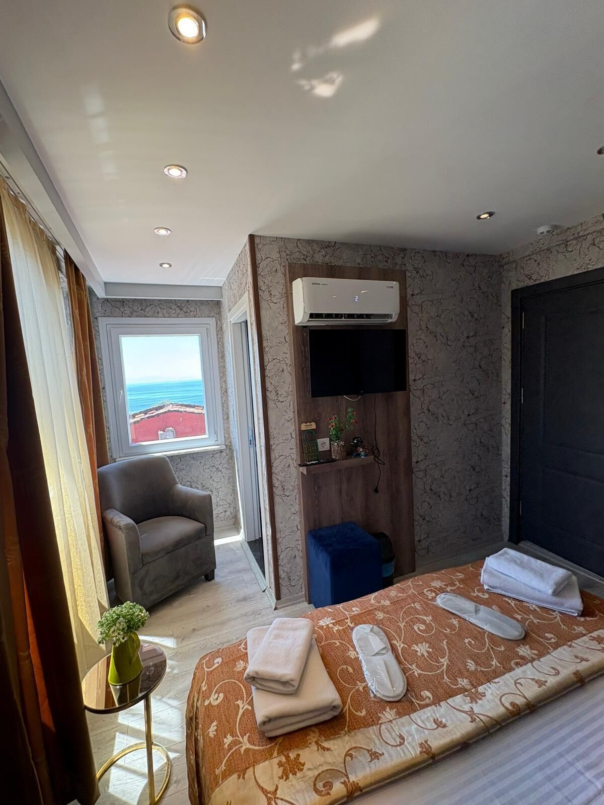 Une chambre d’hôtel avec un lit, une chaise, un appareil de climatisation, une fenêtre donnant sur un beau paysage et une petite table avec un vase.