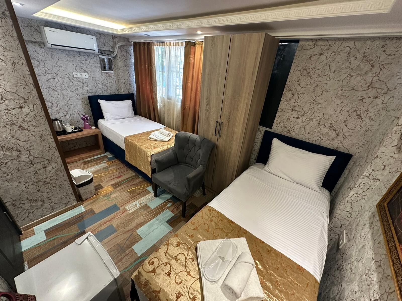 A hotel room with two single beds, a desk with a chair, a small table with a lamp, and a wardrobe.