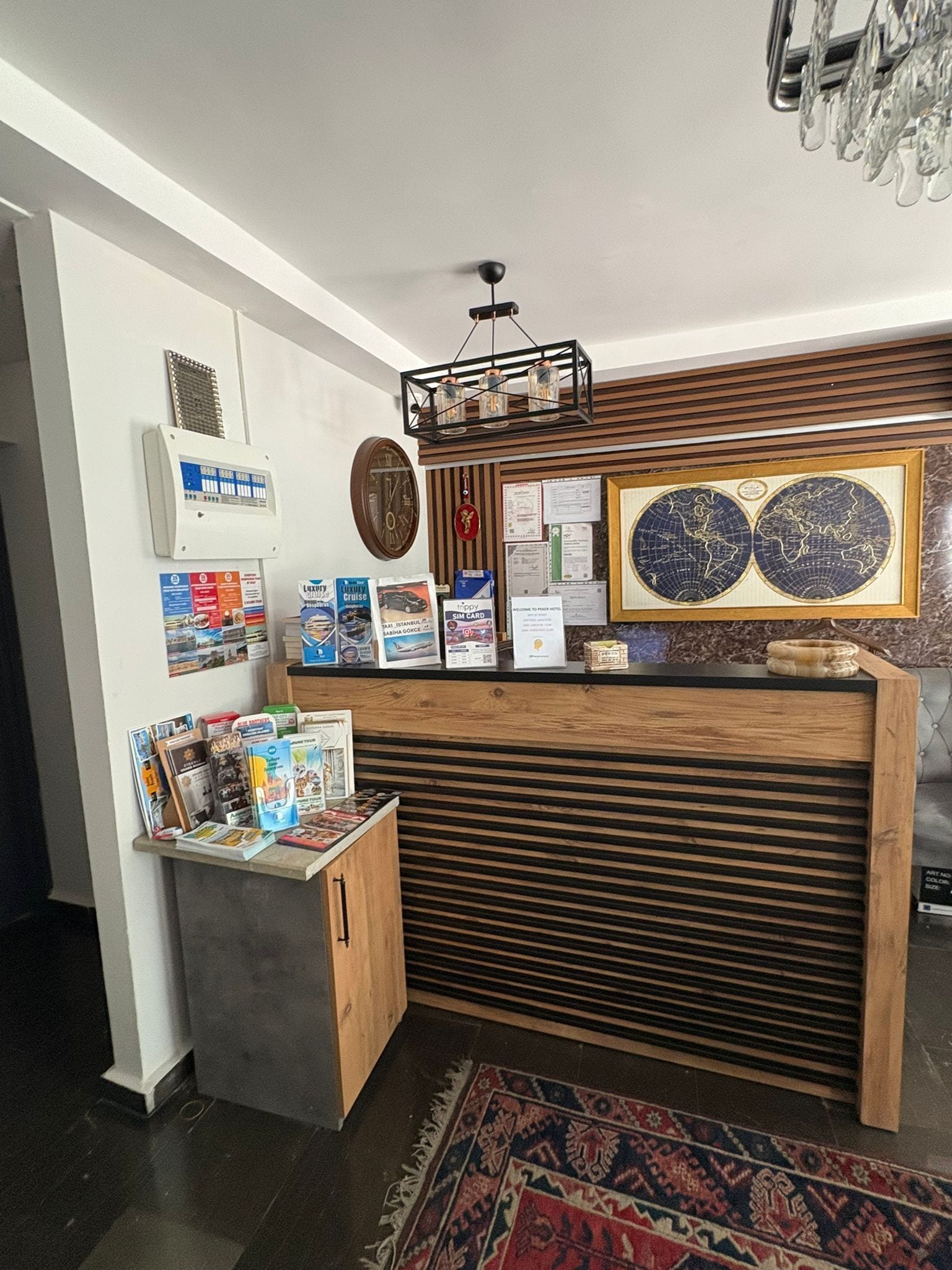 Un mostrador de recepción con detalles de madera, un refrigerador pequeño y una alfombra estampada. En la pared hay mapas enmarcados y varios carteles informativos.