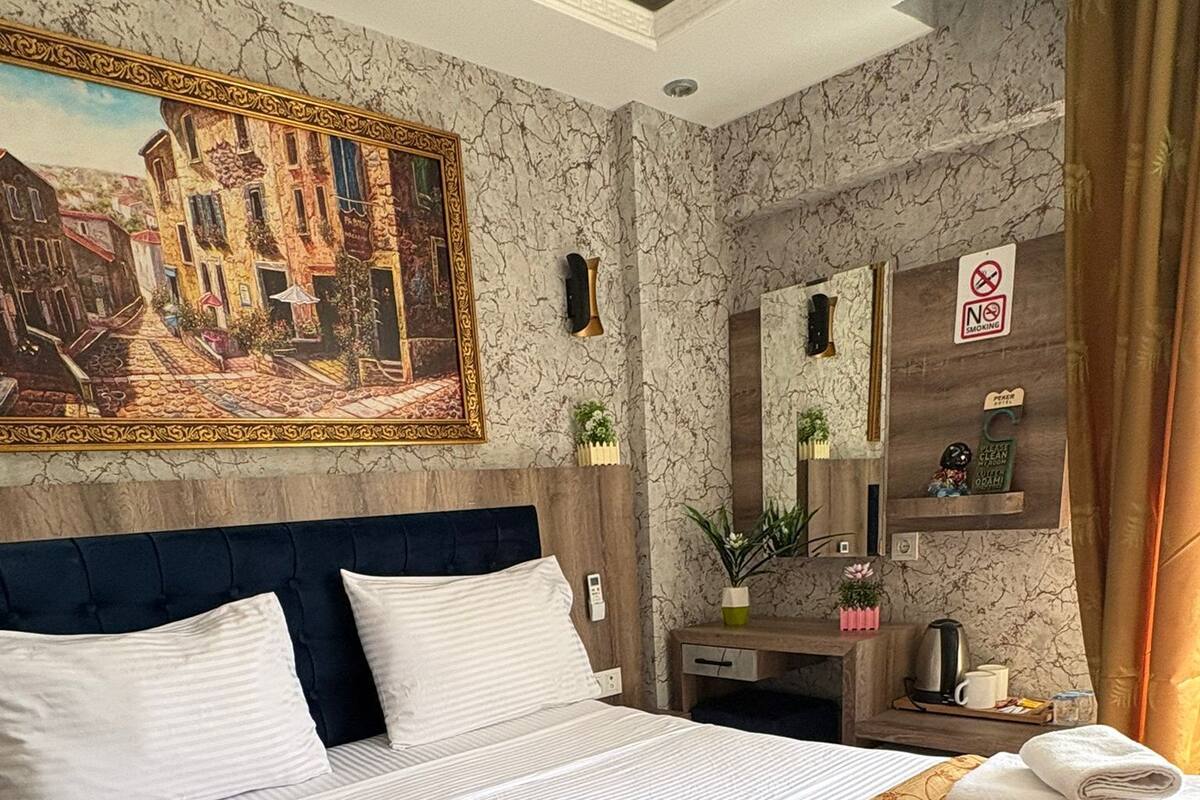 Habitación de hotel con una cama grande, un cuadro en la pared, una mesita de noche de madera, un espejo y un cartel de 'No Fumar'.