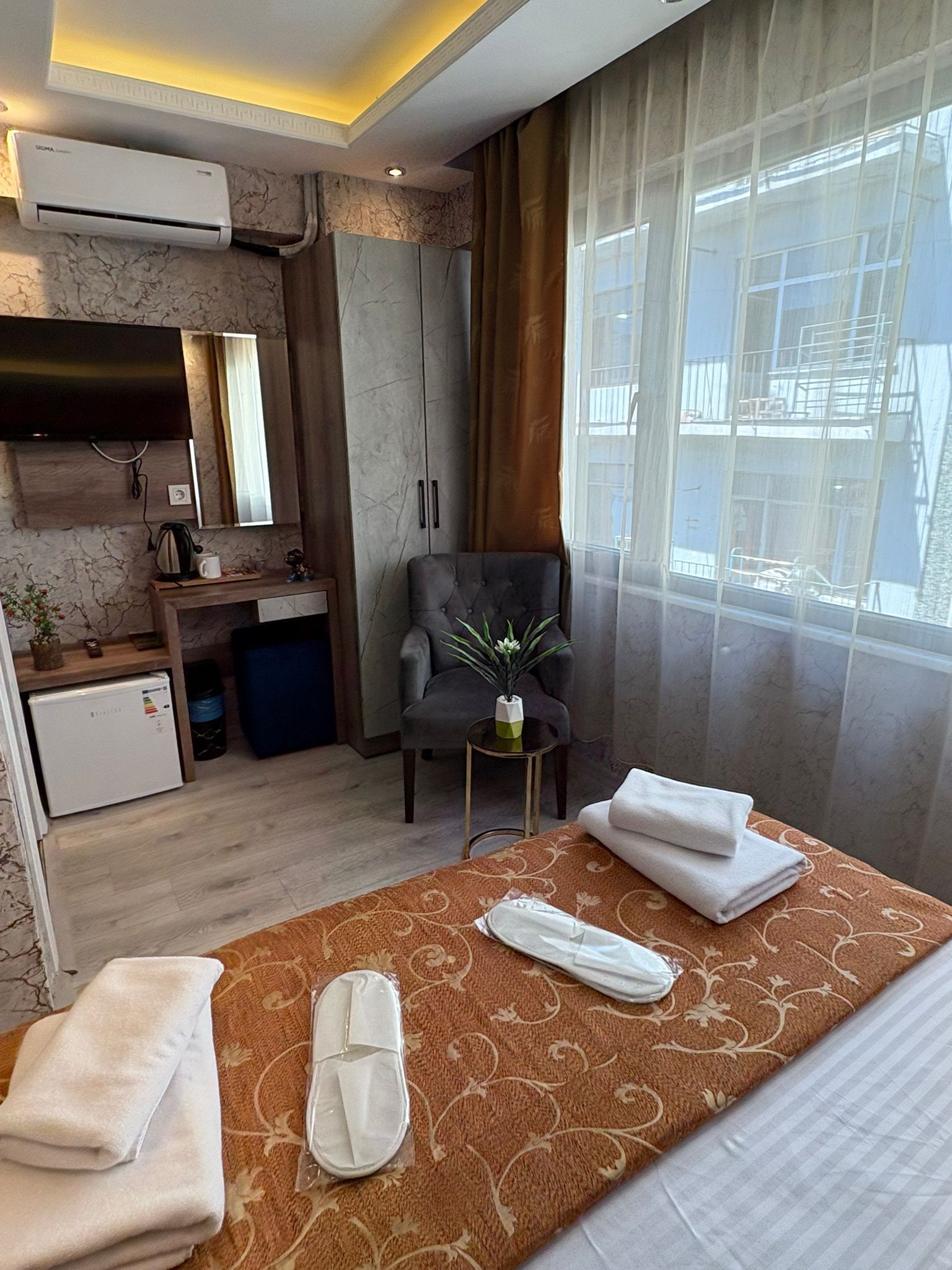 Una habitación de hotel con cama, una silla, una mesita con una planta, un escritorio con computadora y una ventana con cortinas.