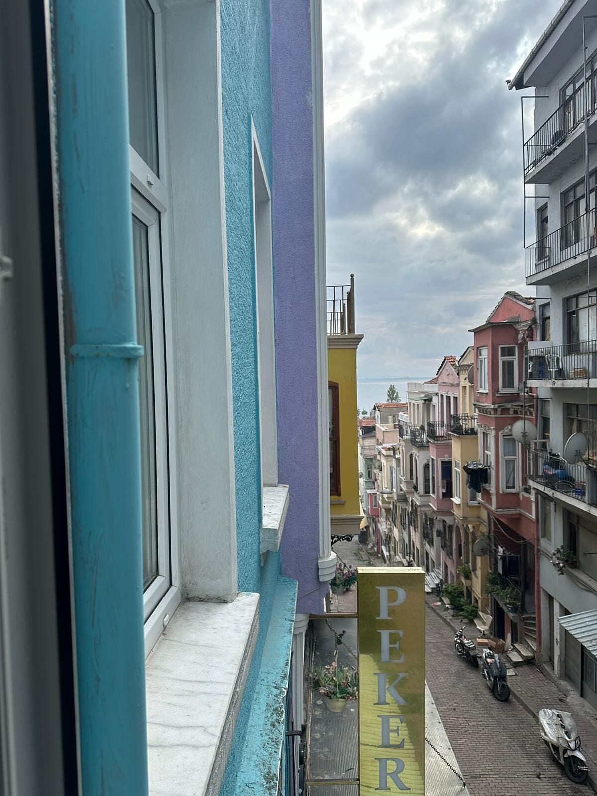 Une rue étroite bordée d’immeubles aux couleurs vives, une enseigne indiquant « PEKER », et des motos garées.