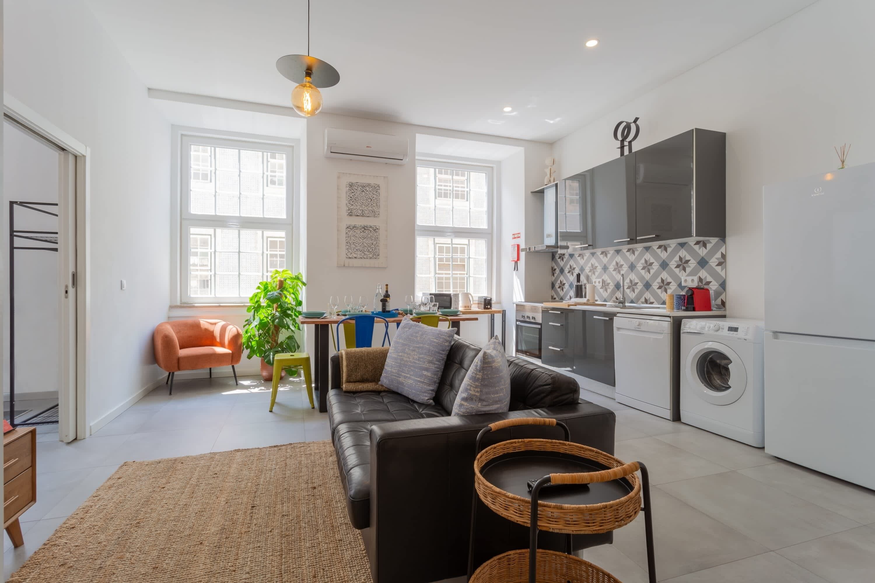 A modern kitchen with a washing machine, a dining area with a sofa, and a large window.