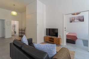 Una sala de estar moderna con un sofá de cuero negro, un televisor sobre un mueble de madera y una puerta que da a un dormitorio con una cama.