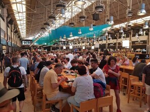 A crowded indoor space with tables and chairs, high ceilings, and hanging lights.