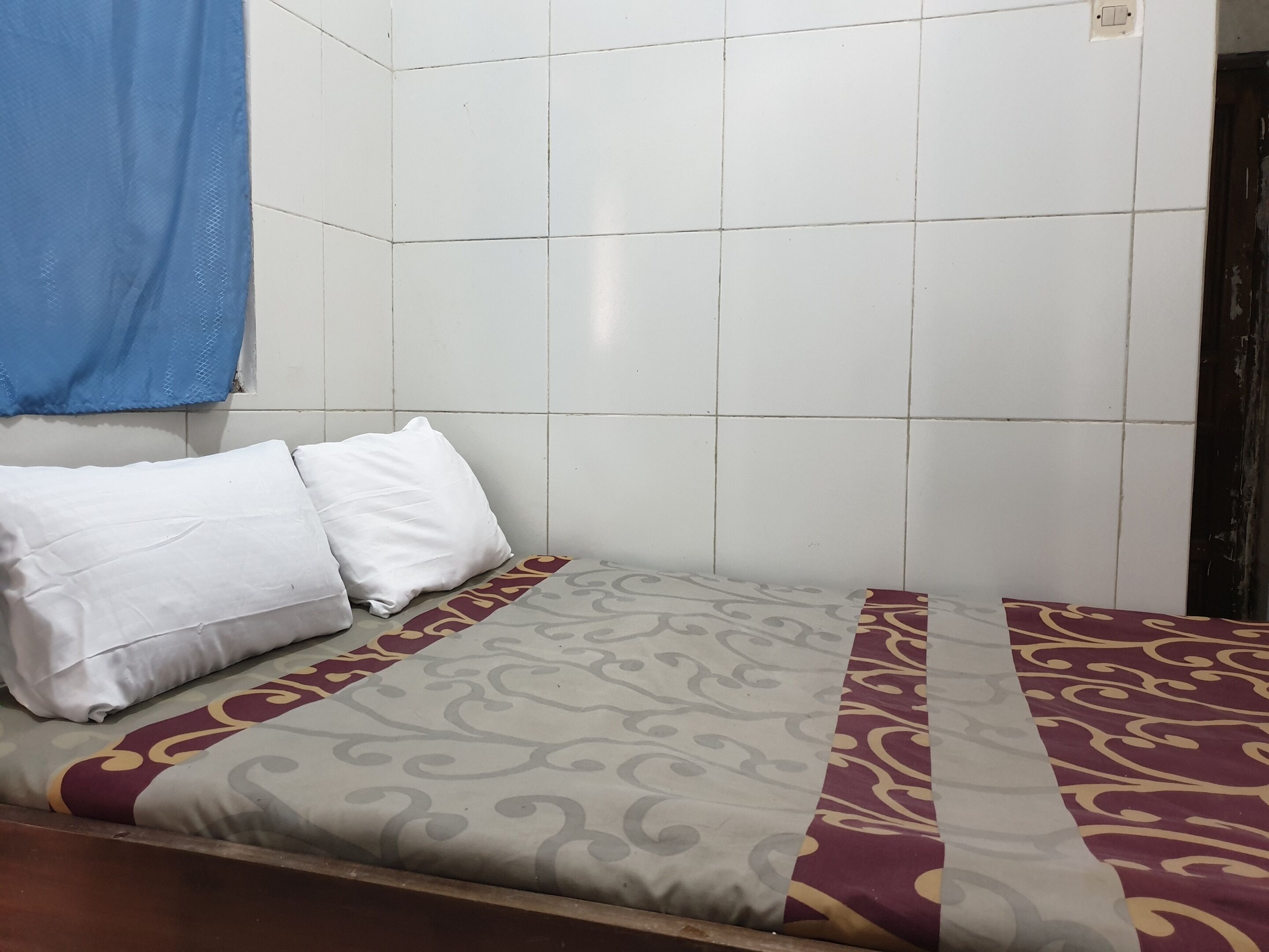 A small, single-bed room with white tiled walls, a patterned bedspread, and two white pillows.