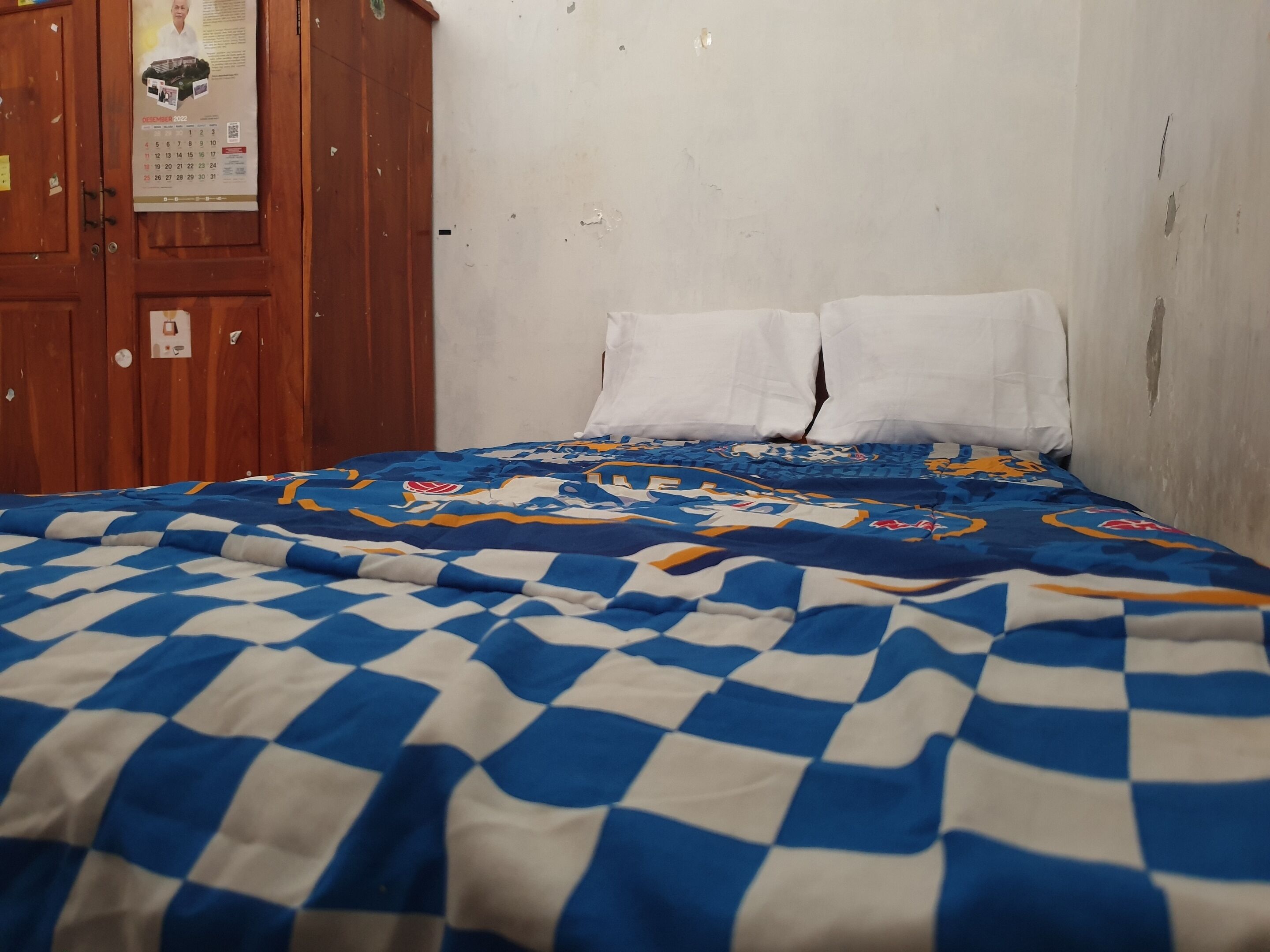 A room with a bed covered in a blue and white checkered blanket, a wooden wardrobe, and a calendar on the wall.