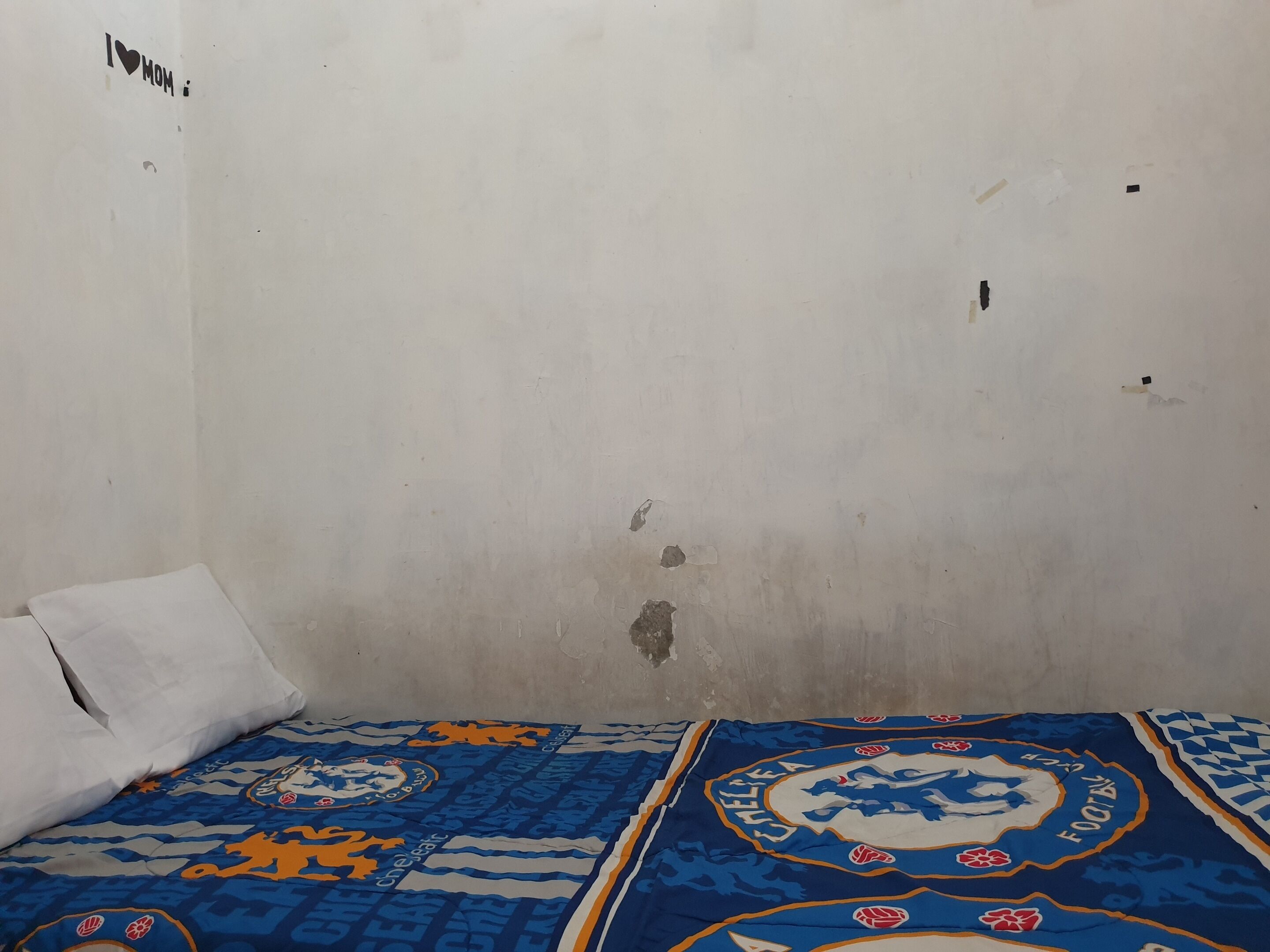 A room with a single bed, a white pillow, and a wall with a 'I ❤️ MOM' sticker.