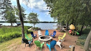 A lakeside outdoor seating area with colorful Adirondack chairs arranged around a central fire pit. - Long Lake North Periwinkle (Phillips)