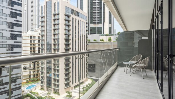 A balcony with a view of high-rise buildings, a swimming pool, and a small seating area with chairs. - Elegant Apartment Amidst Vibrant Area with Balcony (Dubai)