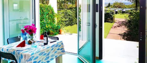 A dining area with a table set for two, a vase with flowers, and a view of a garden through a glass door.
