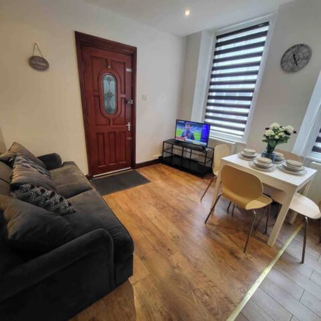 A living room with a dark-colored sofa, a television on a stand, a dining table with chairs, and a wooden floor.