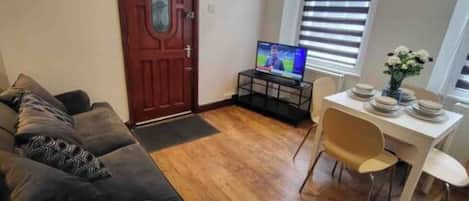 A living room with a dark-colored sofa, a television on a stand, a dining table with chairs, and a wooden floor.