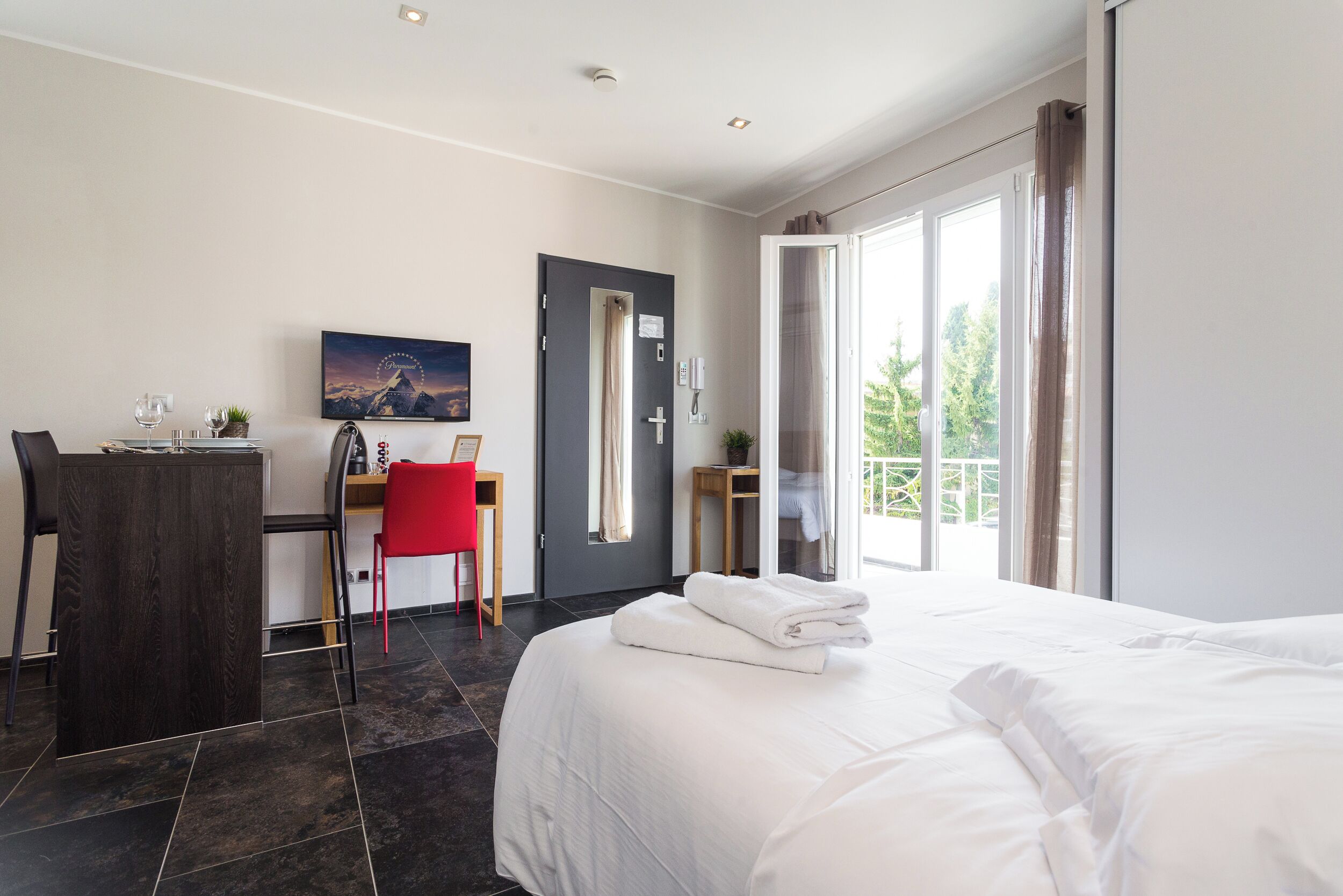 A hotel room with a bed, a desk with a chair, a small table with a plant, and a door leading to a balcony.