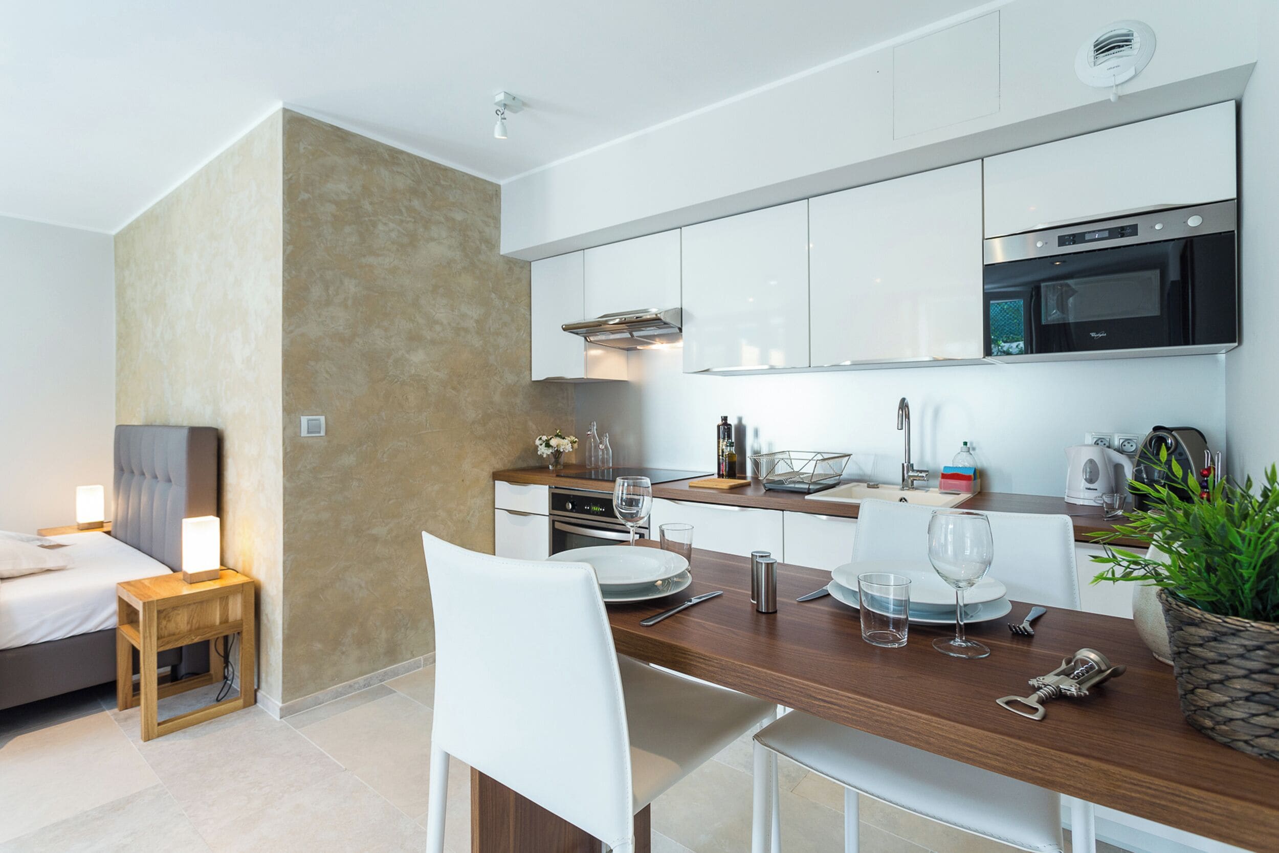 A modern kitchen with white cabinets, a microwave, and a built-in oven. There is a dining table with chairs, a small side table, and a potted plant.