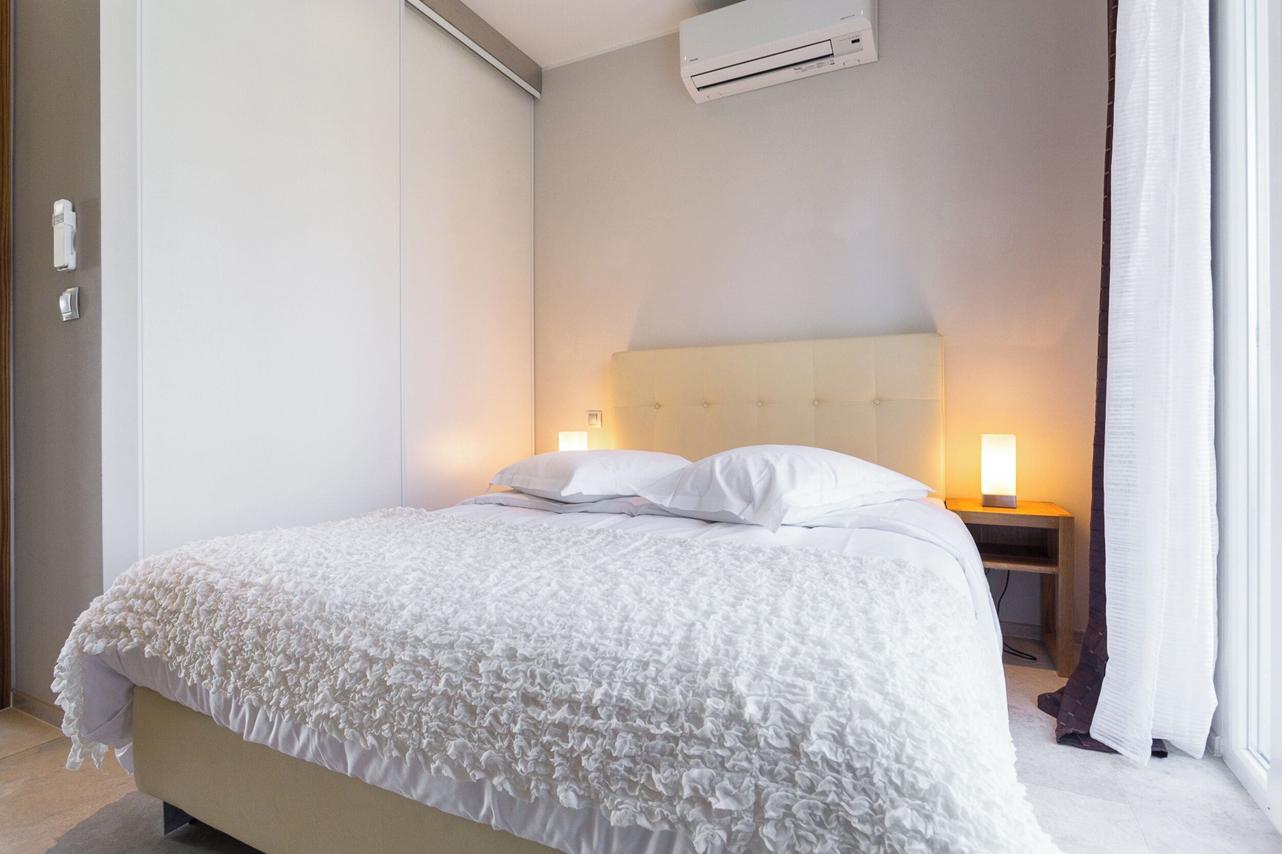 A neatly made bed with a white duvet and pillows, a bedside table with a lamp, and an air conditioning unit on the wall.