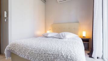 A neatly made bed with a white duvet and pillows, a bedside table with a lamp, and an air conditioning unit on the wall.