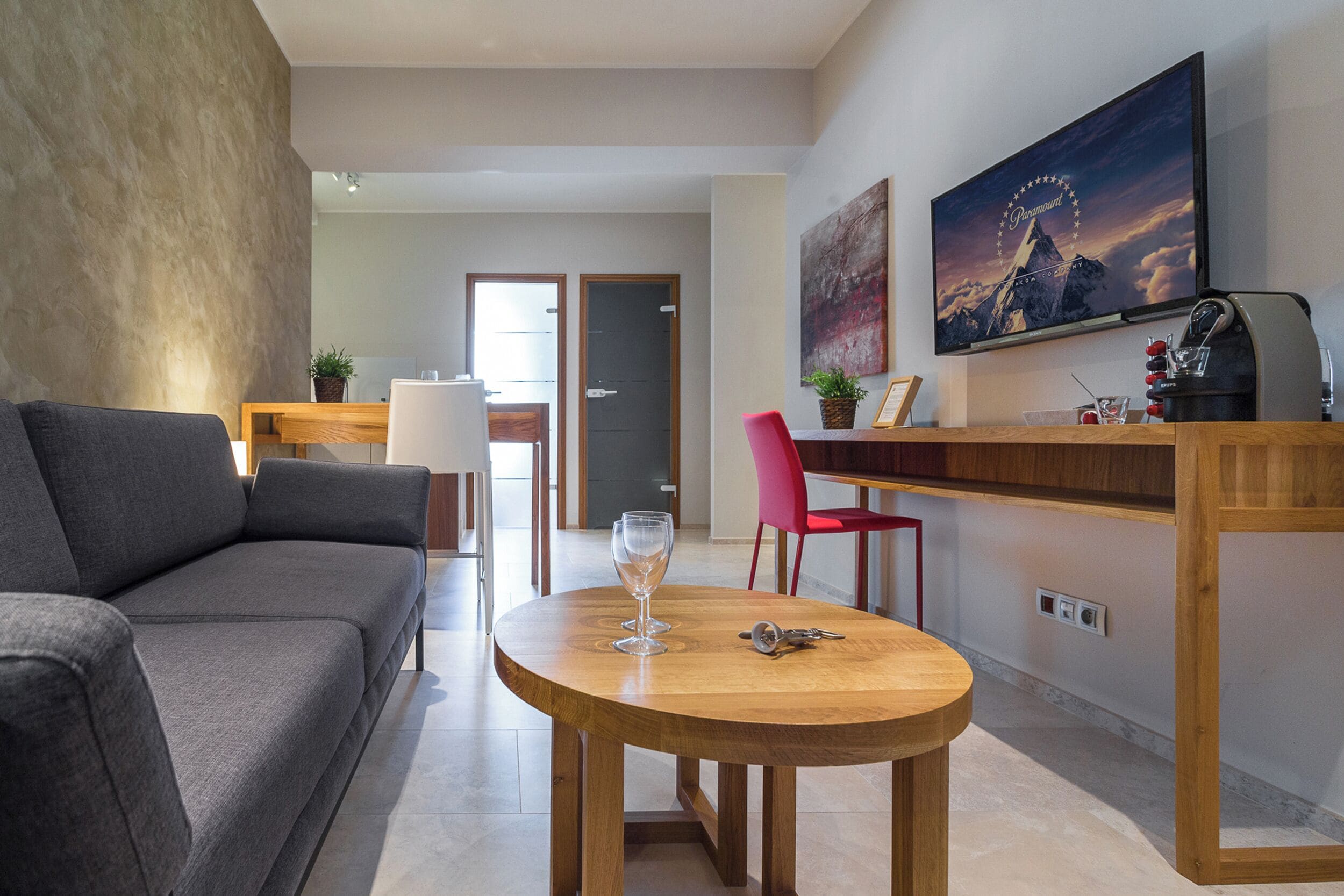 Una sala de estar moderna con un sofá gris, una mesa de centro de madera con una copa de vino, una silla roja y un televisor en la pared que muestra un paisaje de montaña.
