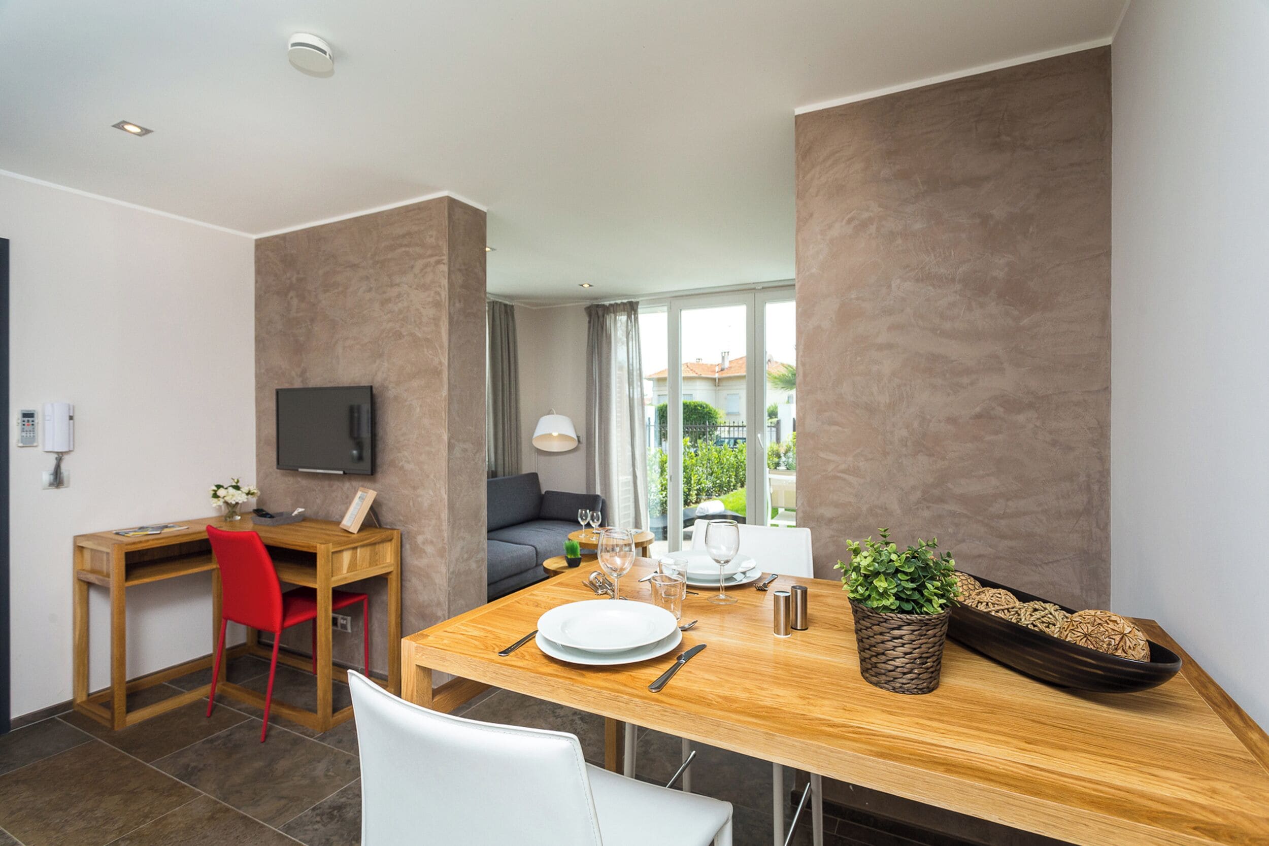 A modern dining area with a wooden table set for two, a red chair, and a white chair. A television is mounted on the wall, and there is a window with a view of greenery outside.