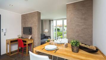 A modern dining area with a wooden table set for two, a red chair, and a white chair. A television is mounted on the wall, and there is a window with a view of greenery outside.