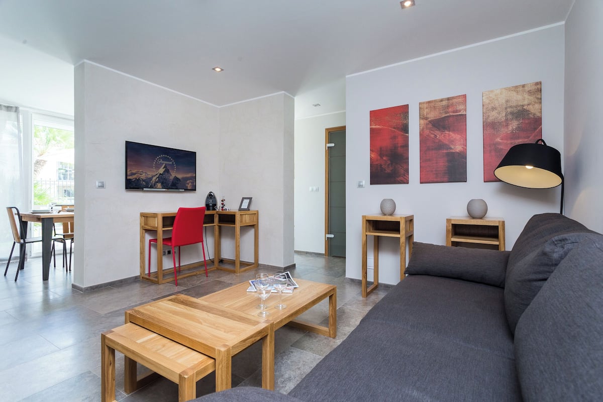 Un salon moderne avec une table basse en bois, un canapé gris et un fauteuil rouge. Trois tableaux ornent le mur et une lampe est posée sur une table d’appoint.