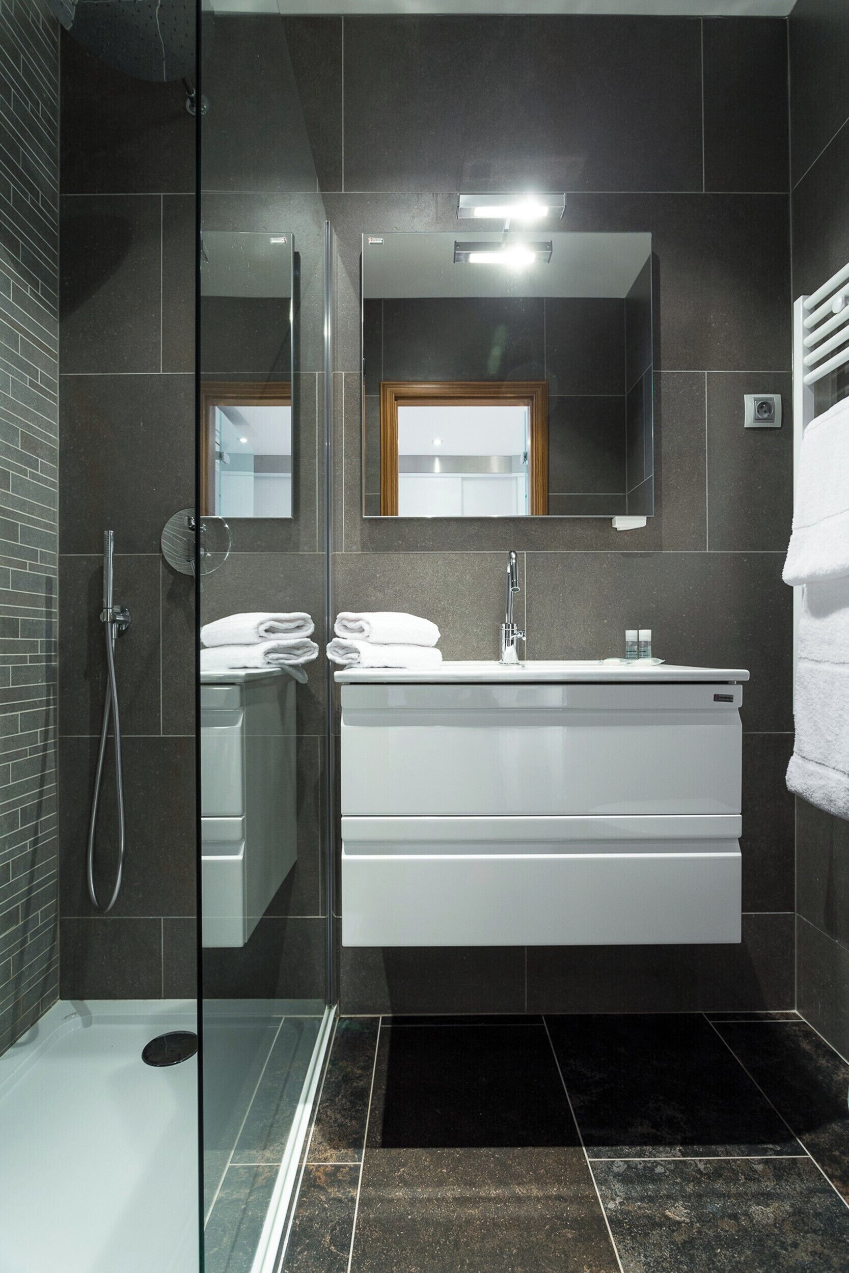A modern bathroom with a glass shower enclosure, a white vanity with a mirror above, and a dark tiled floor.