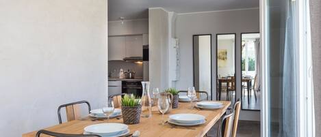Un coin repas avec une table en bois dressée pour quatre personnes, entourée de chaises. On aperçoit une cuisine au fond, et une porte-fenêtre coulissante qui donne sur un espace extérieur.