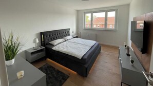A modern bedroom with a large bed, bedside tables, a dresser, a TV, and a window with a view of buildings. - 2 Bedroom Apartment in Magdeburg (Magdeburg)