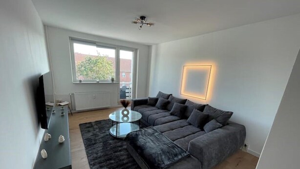 A modern living room with a sectional sofa, a glass coffee table, a flat-screen TV, and a large window providing natural light. - 2 Bedroom Apartment in Magdeburg (Magdeburg)